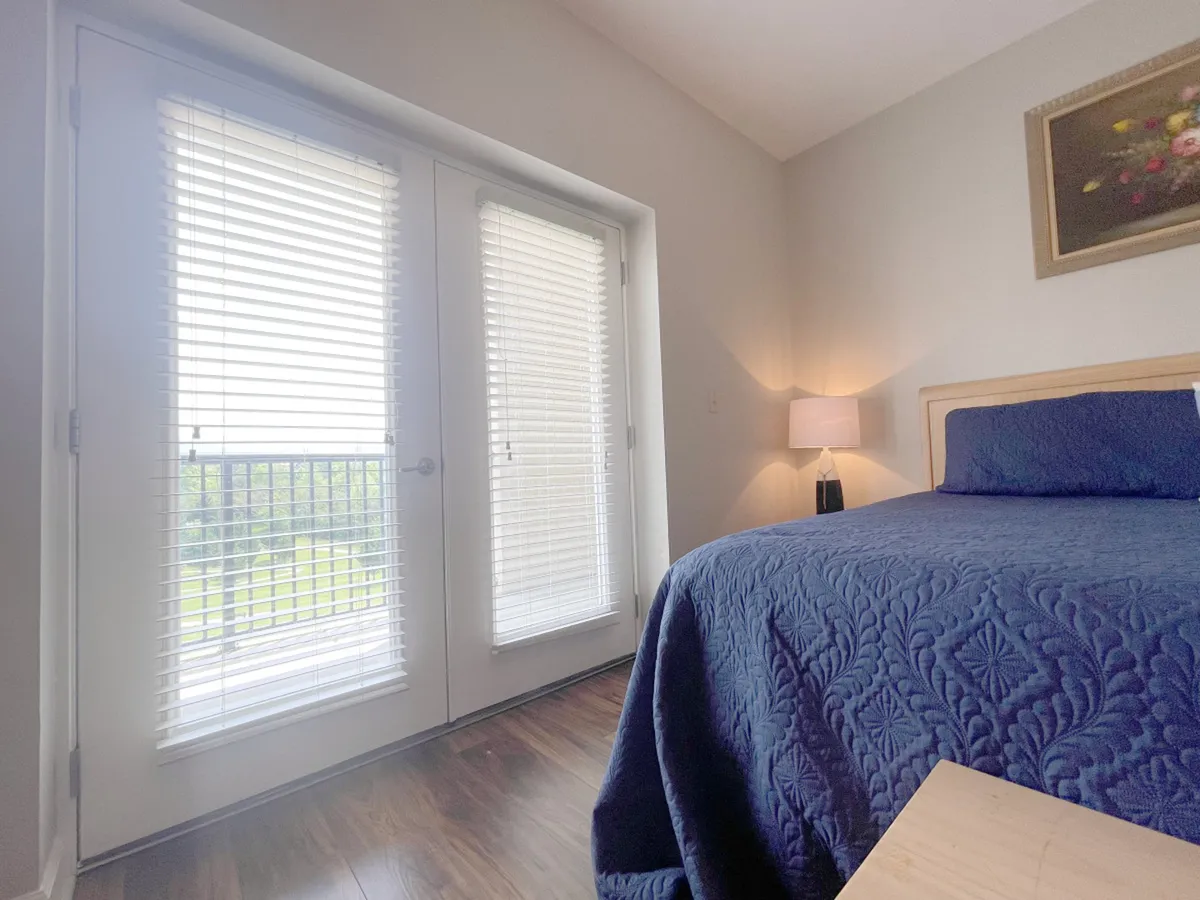 A serene bedroom with a king-sized bed adorned with a blue quilt, flanked by a lamp and decorative painting. Sliding glass doors let in natural light, creating a peaceful retreat perfect for unwinding after an adventurous day.