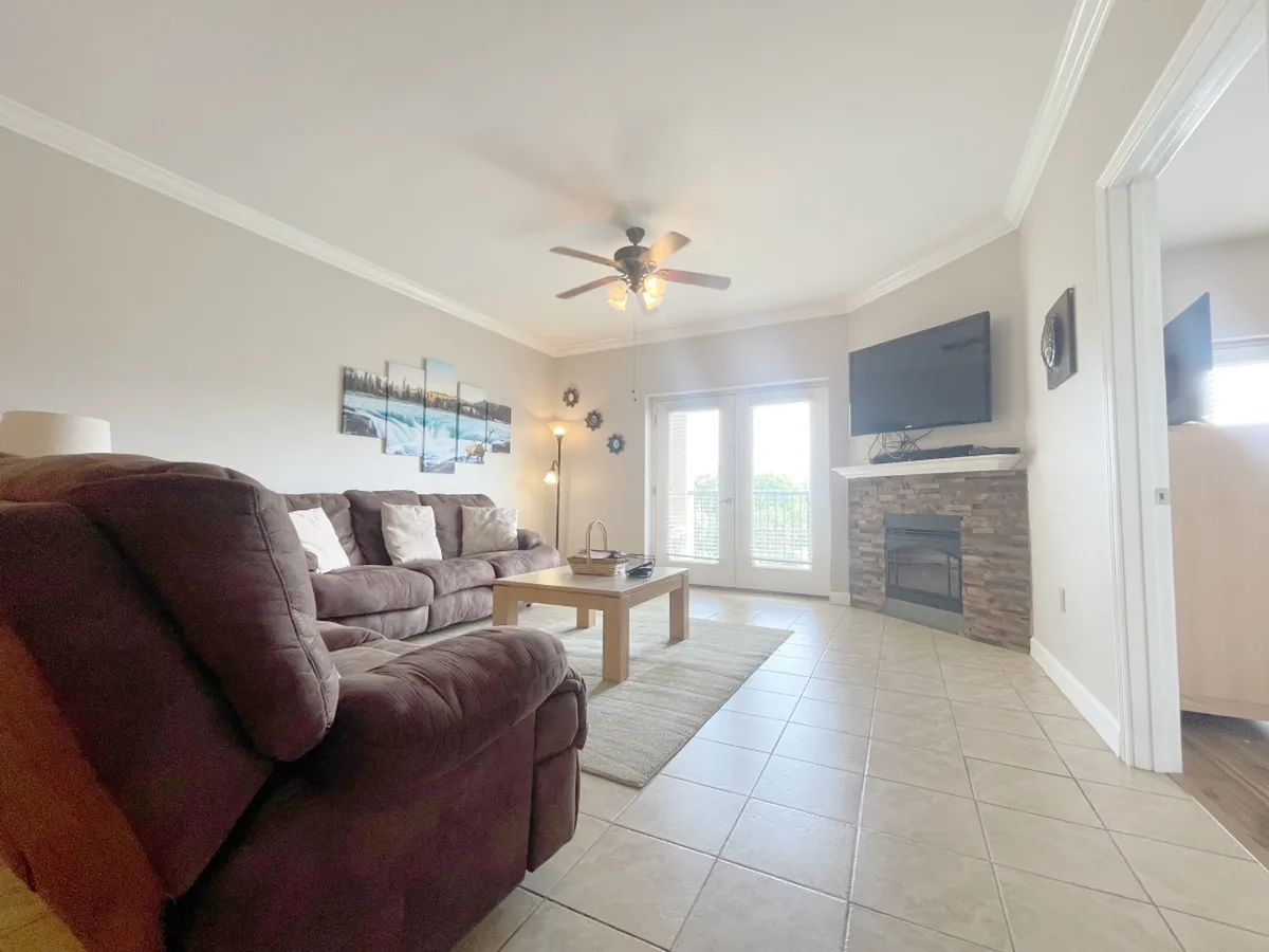 A welcoming living room with a comfortable brown sofa, wooden coffee table, fireplace, and a large flat screen TV mounted on the wall. Decorative artwork and natural light from the windows create a cozy, inviting environment.