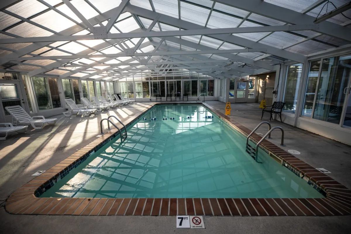 The image showcases a bright, spacious indoor swimming pool area with lounge chairs arranged along the perimeter and large windows letting in natural light. The pool deck is well maintained and inviting, perfect for year-round swimming and relaxation.