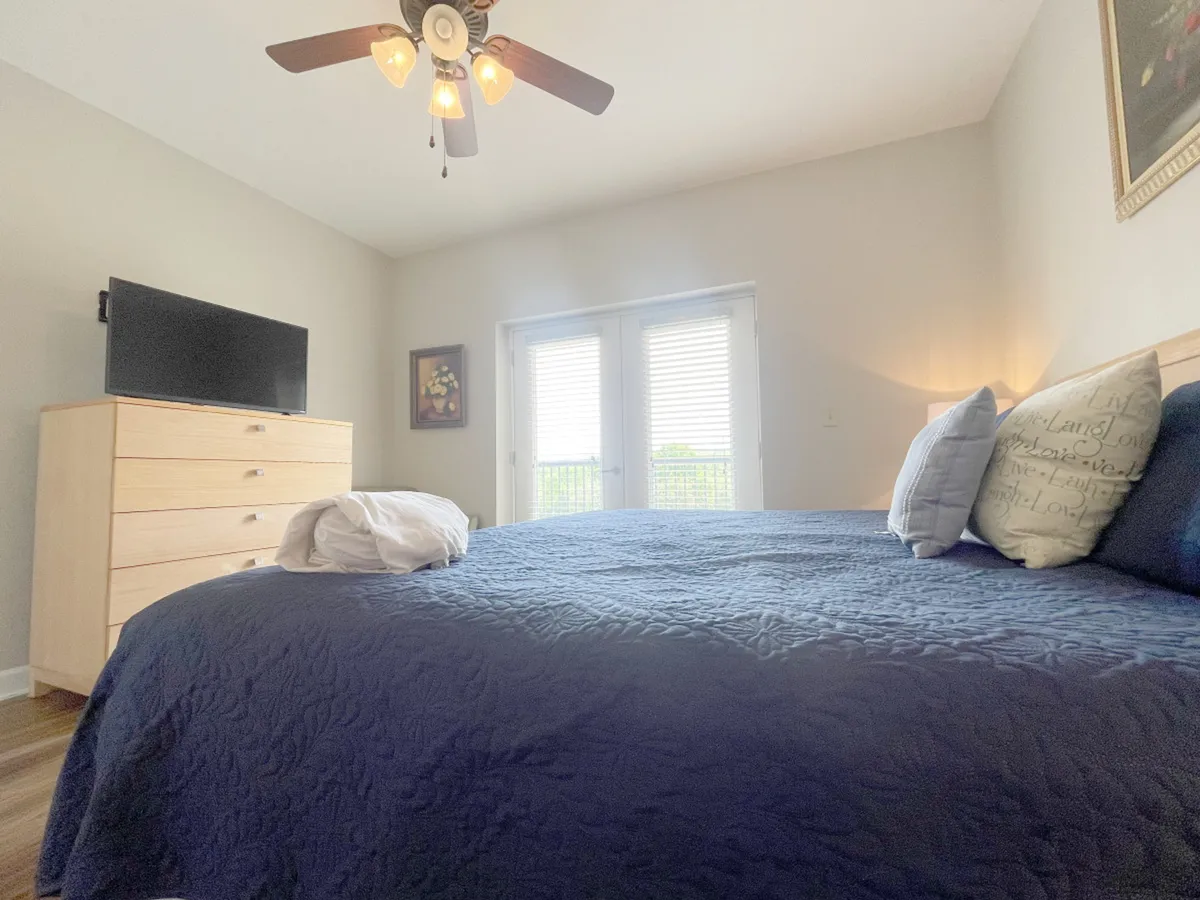 A spacious bedroom featuring a bed with a navy blue quilt, a wall-mounted television, and double doors leading outside. Decorative artwork and a tranquil mood reinforce the restful retreat vibe.